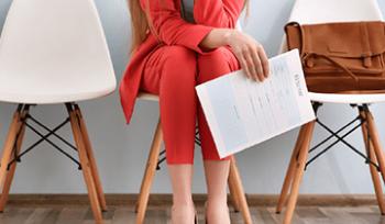 A person dressed in professional attire sits in a chair with a briefcase and a resume in their hand